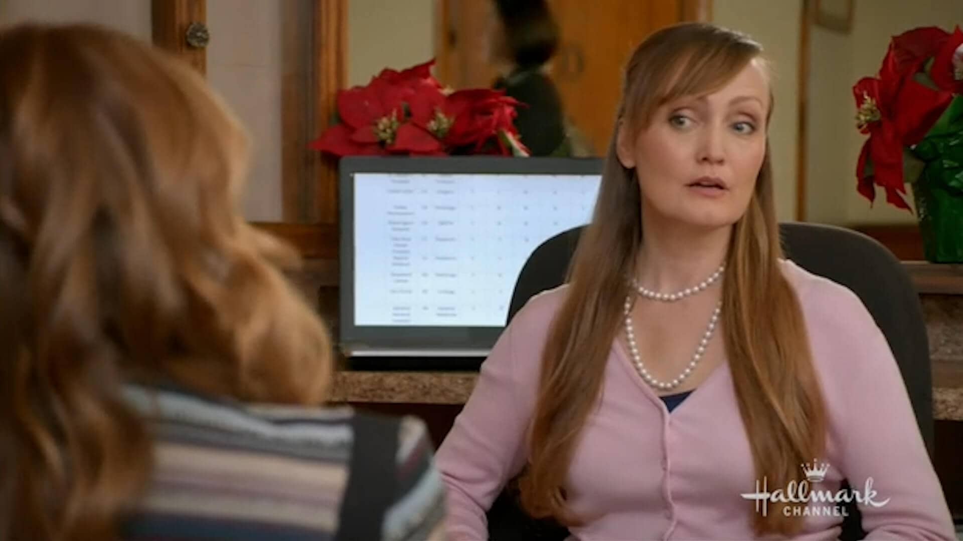Still from Christmas Under Wraps. Julie-Anne sits at a receptionist desk and listens inquisitively to a girl with red hair.
